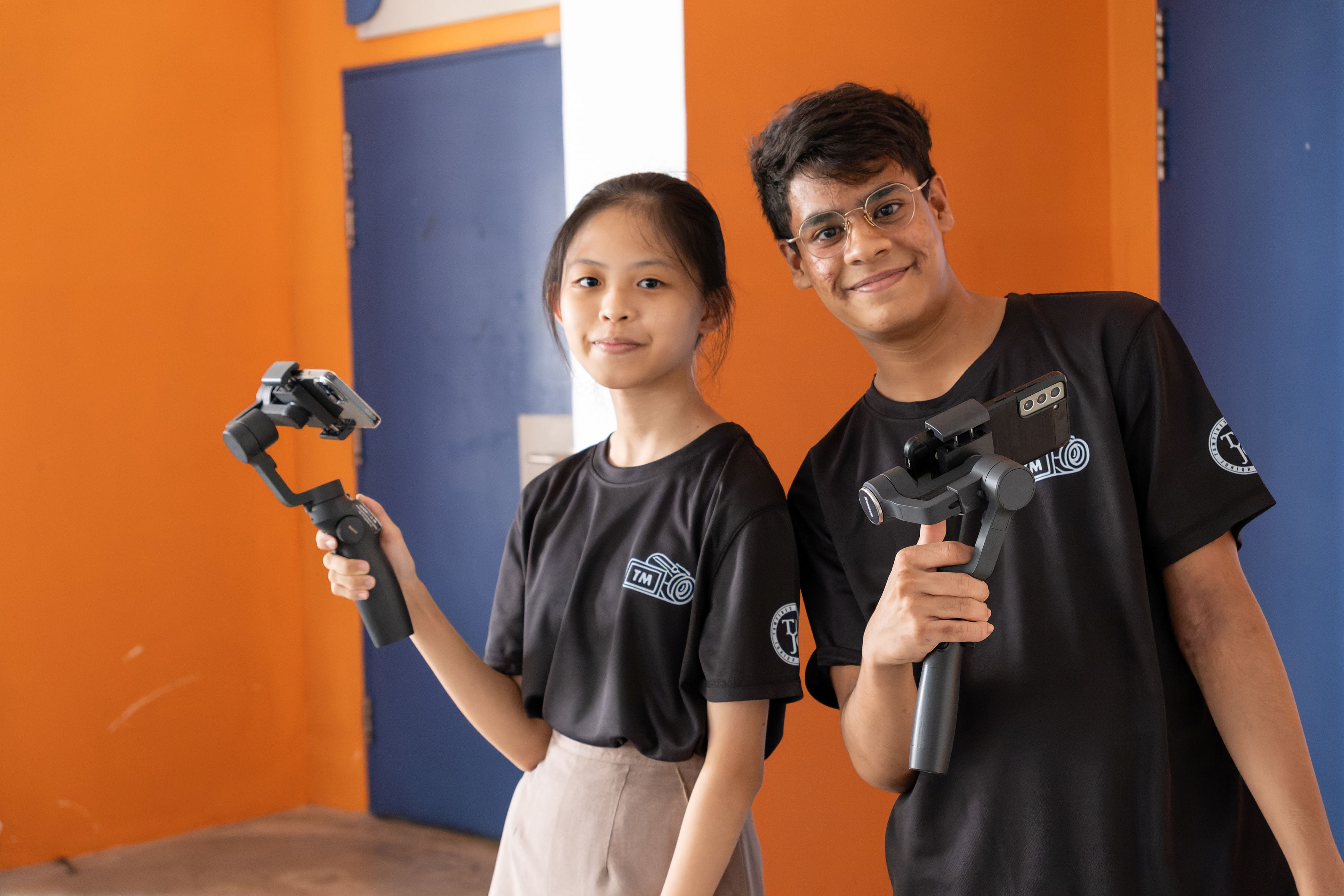 Two students in black club T-shirts stand against an orange wall, each holding a smartphone gimbal stabiliser.